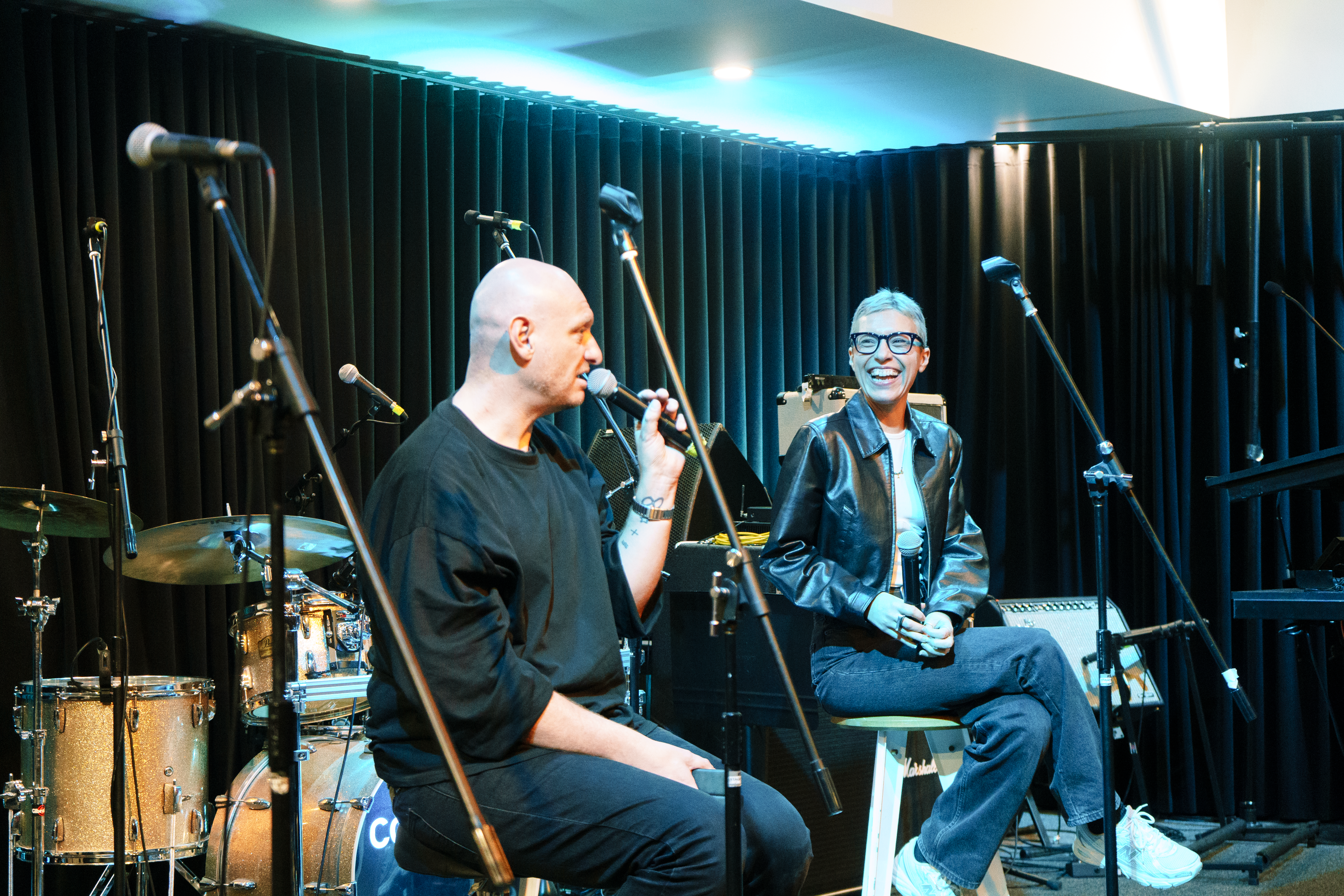 Two people sitting on a stage speaking interview style with several microphones and a drum kit behind them